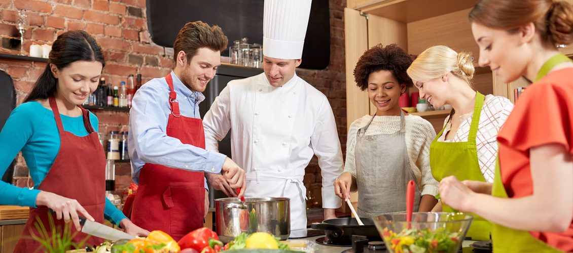 happy friends and chef cook cooking in kitchen cooking class, culinary, food and people concept - happy group of friends and male chef cook cooking in kitchen